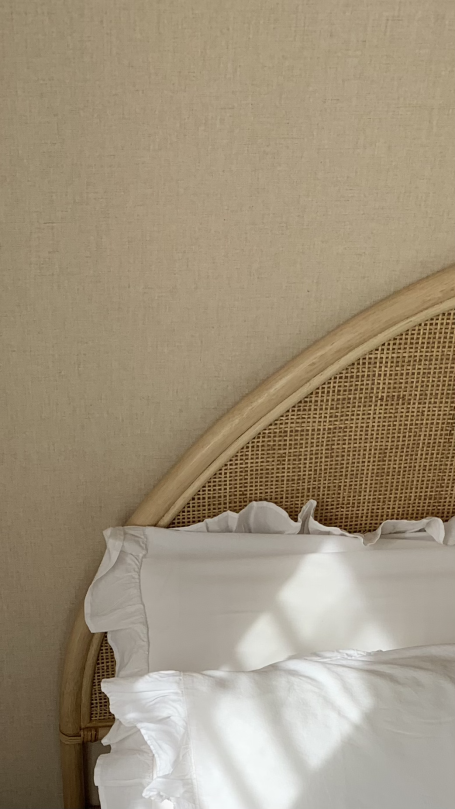 Cabecero de cama con tejido de mimbre y sábanas blancas, iluminado por la luz del sol.